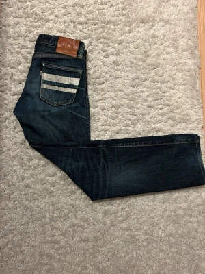 Momotaro selvage jeans - Snygga mörkblå jeans med raka ben och coola vita ränder på ena bakfickan. Jeansen har klassisk femficksdesign och en brun läderpatch vid linningen. Perfekta för en avslappnad streetstil.