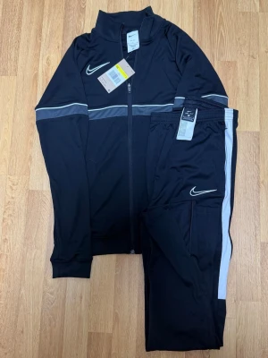 Säljer en Nike Dri-FIT träningsdress Black – Top S / Pants M - Träningsdressen är helt ny och oanvänd. Inga skador eller slitage.  Bekväm och funktionell Dri-FIT träningsdress som passar perfekt till både träning och vardag.  Size: Tröja S / Byxa M 🏷️ Rabatt vid köp av flera plagg!