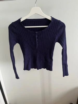 Mörkblå ribbstickad tröja - Snygg mörkblå ribbstickad tröja med rund halsringning och knappdetaljer framtill. Tröjan har långa ärmar och är figurnära med stretchigt material. Perfekt att matcha med jeans eller kjol för en trendig look.
