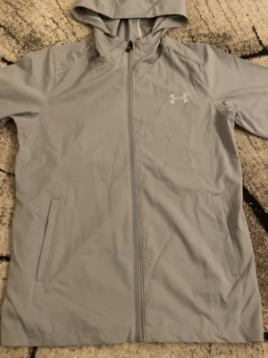 Ljusgrå vindjacka från Under Armour - Snygg ljusgrå vindjacka från Under Armour med huva och dragkedja hela vägen. Jackan har två sidofickor och en diskret logga på bröstet. Perfekt för blåsiga dagar och enkel att matcha med olika outfits.