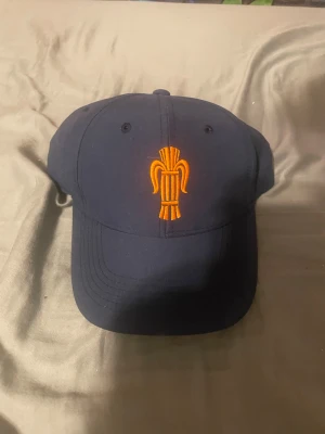 Mörkblå keps med orange brodyr - Snygg mörkblå keps från Kappa med en unik orange brodyr framtill. Kepsen har böjd skärm och justerbar rem baktill för perfekt passform. Tillverkad i bomullsmaterial som andas. Perfekt accessoar för en cool och avslappnad stil.