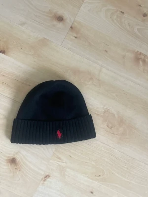 Svart mössa från Polo Ralph Lauren - Säljer en svart ribbstickad mössa från Polo Ralph Lauren med klassisk röd logga broderad framtill. Mössan är i mjukt material och har uppvikt kant för extra stilpoäng. Perfekt accessoar för kyliga dagar.