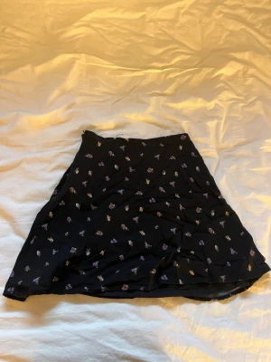 Svart blommig kortkjol - Svart kortkjol med små blommiga mönster i lila och vitt. Kjolen har en lätt utsvängd form och är tillverkad i ett tunt, mjukt material som ger ett snyggt fall. Perfekt för dig som gillar en feminin och trendig stil.