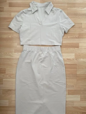 Beige tvådelat set med kjol och topp - Snyggt tvådelat set i beige med en croppad topp med krage och v-ringning samt en högmidjad, vadlång kjol med slits bak. Setet är stilrent och passar perfekt för en trendig look. Materialet ser ut att vara mjukt och stretchigt, vilket ger en bekväm passform.