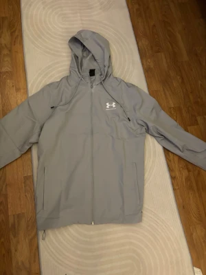 Ljusgrå vindjacka från Under Armour - Snygg ljusgrå vindjacka från Under Armour med huva och dragkedja framtill. Jackan har två sidofickor med dragkedja och justerbar nederkant. Tillverkad i lätt och funktionellt material, perfekt för blåsiga dagar.