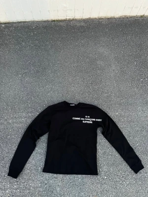 Svart långärmad topp Supreme x Comme des Garçons - Svart långärmad topp från Supreme x Comme des Garçons med vit text på bröstet. Klassisk crewneck och mjukt bomullsmaterial. Perfekt för dig som gillar streetwear och vill sticka ut med en ikonisk collab.