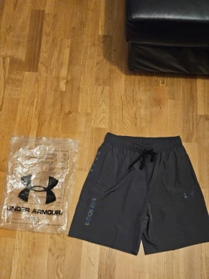 Svarta träningsshorts från Under Armour - Snygga svarta träningsshorts från Under Armour med elastisk midja och snörning. Shortsen har logga och texten 'ARMOUR' i vitt på ena benet samt praktiska sidofickor. Perfekta för gymmet eller löprundan.