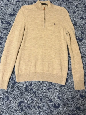 Morris halt zip - Snygg beige stickad tröja från Morris med halv dragkedja och diskret logga broderad på bröstet. Tröjan har långa ärmar, ribbade muddar och är tillverkad i mjukt material som känns skönt mot huden. Perfekt för lager på lager och stilrena outfits.