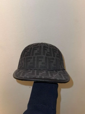 Svart keps från Fendi med FF-mönster - Snygg svart keps från Fendi med det klassiska FF-monogrammet över hela kepsen. Kepsen har böjd skärm med läderdetalj och är tillverkad i tyg med exklusiv känsla. Möts upp Göteborg 