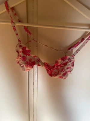 Rosa blommig bikini med volang - Supersöt bikini med volangdetaljer och blommigt mönster i rosa och rött på vit botten. ANVÄND 2-3 GÅNGER. Överdelen har bygel och smala axelband. Underdelen har matchande volangkant. Perfekt för strandhäng i sommar!