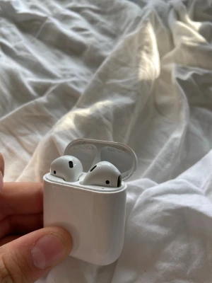 Apple AirPods med laddningsetui - Säljer ett par vita Apple AirPods med tillhörande laddningsetui. Trådlösa hörlurar i stilren och kompakt design, perfekt för musik, samtal och poddar. Laddningsetuiet är i blankt plastmaterial och hörlurarna har en smidig passform för öronen.