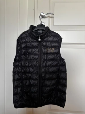 Svart väst från EA7 Emporio Armani - Snygg svart väst från EA7 Emporio Armani med guldfärgad logga på bröstet. Västen har hög krage, dragkedja framtill och quiltad design. Perfekt för lager-på-lager. Västen har även ett märke från när jag råkade skära upp den men det är inget som syns när den används 