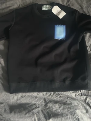 Svart sweatshirt från Casablanca - Cool svart sweatshirt från Casablanca med blått grafiskt tryck och texten 'Casa Blanca' både fram och bak. Tröjan har rund halsringning och ribbade muddar vid ärmslut och nederkant. Perfekt för en stilren och modern look.säljer denna för den var för liten för mig 