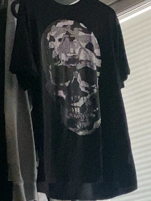 Svart t-shirt med dödskalletryck - Cool svart t-shirt med stort dödskalletryck i camouflagemönster på framsidan. Perfekt för dig som gillar edgy och streetwear-stil. Klassisk passform och rund halsringning.