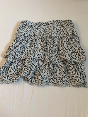 Vit blommig volangkjol från Lindex!! - Supersöt vit kjol från Lindex med svart blommigt mönster och flera volanger. Kjolen är kort och har en luftig känsla, perfekt för varma dagar. Resår i midjan för skön passform. Passar dig som gillar en romantisk och trendig stil.