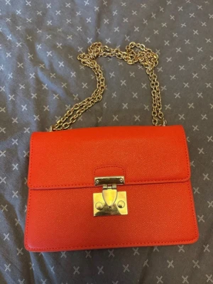 Orange axelväska med guldkedja - Snygg orange axelväska i boxig modell med guldfärgad kedjerem och metallspänne framtill. Väskan har en stilren design och är tillverkad i ett strukturerat konstlädermaterial. Perfekt för dig som vill sticka ut med en färgklick i din outfit.
