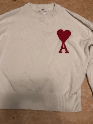 Vit stickad tröja från Ami - Säljer en vit stickad tröja från Ami Alexandre Mattiussi med röd hjärtlogga och bokstaven A på bröstet. Tröjan har rund halsringning, ribbade muddar och är gjord i mjukt material. Perfekt för dig som gillar stilrena plagg med en twist.