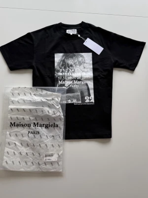 Svart Maison Margiela t-shirt med tryck - Cool svart t-shirt från Maison Margiela med ett stort grafiskt tryck framtill i vitt och grått. Klassisk passform med rund hals och korta ärmar. Perfekt för dig som gillar streetstyle och exklusiva märken. Strl:S✅Pris kan diskuteras!💸 Hör av dig om du har några funderingar!👍Aldrig använd!🤩