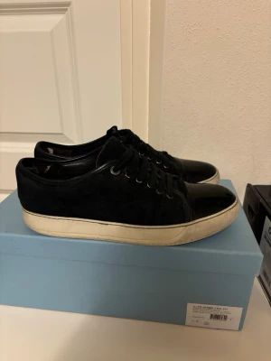  Lanvin  - Snygga svarta sneakers från Lanvin med ovandel i mocka och detaljer i blankt lackläder. Skorna har vita, tjocka gummisulor och svarta skosnören. Perfekta för dig som gillar stilrena och exklusiva sneakers med en twist. Sitter perfekt för storlek 42 passar även dem som har 41