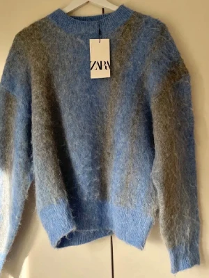 Blå  stickad tröja Zara - Supermjuk stickad tröja från Zara i blått och grått med oversized passform. Tröjan har rund halsringning, långa ärmar och ribbstickade muddar. Materialet är fluffigt och ger en mysig känsla, perfekt för kyliga dagar.