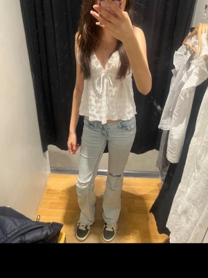 Vit spetsblus med knyt framtill - Söt vit blus med spetsdetaljer och v-ringning. Toppen har ett knyt framtill och är ärmlös, vilket ger en luftig och somrig känsla. Perfekt att matcha med jeans eller kjol för en trendig look.