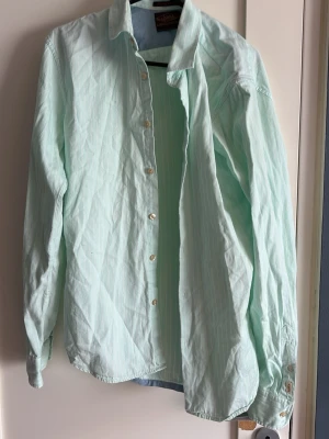 Ljusgrön skjorta från Scotch & Soda - Snygg ljusgrön skjorta från Scotch & Soda med tunna vita ränder. Klassisk krage, långa ärmar och knäppning framtill. Tillverkad i ett lätt bomullstyg som känns fräscht och passar perfekt till många olika stilar.