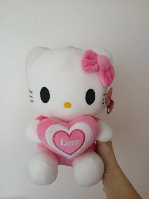 Hello Kitty gosedjur med hjärta - Supergulligt Hello Kitty gosedjur i vitt och rosa plysch. Katten har en rosa rosett på huvudet och håller ett hjärta med texten 'Love' broderat på. Perfekt som present eller dekoration. Originaletikett sitter kvar.
