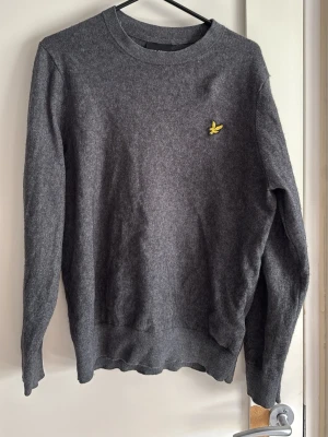 Mörkgrå tröja från Lyle & Scott - Mörkgrå rundhalsad tröja från Lyle & Scott med den klassiska gula örnloggan broderad på bröstet. Tröjan har långa ärmar och ribbade muddar vid ärmslut och nederkant. Perfekt för en stilren och enkel look. Funkar som S