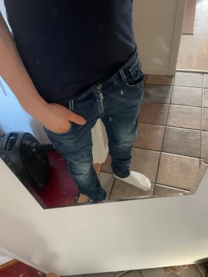 Blå Replay jeans med slim fit - Säljer ett par snygga blå jeans från Replay med slim fit passform. Jeansen har klassisk femficksdesign, slitningar och ljusare partier för en cool, använd look. Tillverkade i mjukt denimtyg med skinnpatch bak i midjan.