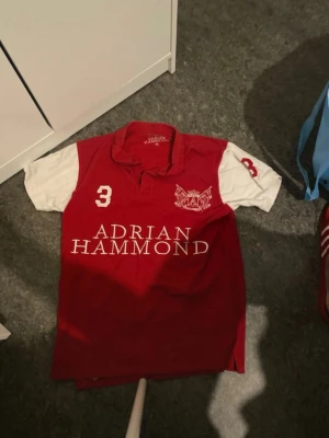 Röd och vit polo tröja Adrian Hammond - Snygg röd polo tröja från Adrian Hammond med vita ärmar och stora siffran 3 på både fram- och baksidan. Klassisk krage och tryckt logga på bröstet. Perfekt för en sportig och avslappnad stil.