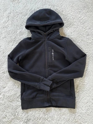 Sail Racing Hoodie - Svart hoodie från Sail Racing med dragkedja framtill och en bröstficka med dragkedja. Broderad logga på bröstet. Den är lite använd och kostar endast 149kr.