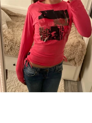 Rosa långärmad topp med tryck - Cool rosa långärmad topp med svart och rosa tryck framtill. Toppen har snörning i sidorna och är figurnära i modellen. Perfekt för dig som gillar streetstyle och vill sticka ut lite extra.