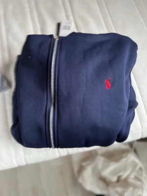 Marinblå hoodie från Polo Ralph Lauren - Snygg marinblå hoodie från Polo Ralph Lauren med dragkedja framtill och den klassiska röda loggan broderad på bröstet. Tillverkad i mjukt bomullsmaterial och har huva med vita snören. Perfekt för en avslappnad och stilren look.