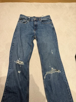 Blå slitna jeans från Zara - Säljer ett par blå jeans från Zara med raka ben och slitningar på båda benen. Jeansen har klassisk femficksdesign, hög midja och är gjorda i denim. Perfekta för en avslappnad och trendig look. Aldrig använda 