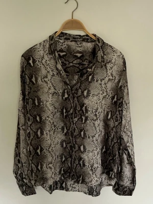 Neo noir långärmad skjorta - Snygg långärmad skjorta med ormmönster i svart, grått och beige. Skjortan har klassisk krage och knappar framtill. Perfekt för dig som vill sticka ut med en trendig och djärv look.