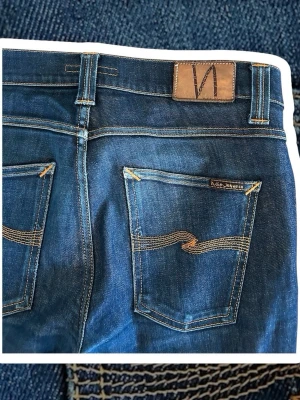  Nudie jeans slim fit W31 L32 - Säljer ett par mörkblå skinny jeans från Nudie Jeans med klassiska kontrastsömmar och signaturbroderi på bakfickorna. Jeansen har fem fickor, låg midja och är tillverkade i slitstarkt denim. Perfekta för en avslappnad och trendig look.
