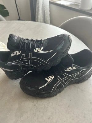 Svarta Asics sneakers med vita detaljer - Snygga svarta sneakers från Asics med vita och grå detaljer samt reflekterande partier. Skorna har en chunky sula med stänk-effekt och klassisk snörning. Perfekta för dig som gillar sportig och modern stil.