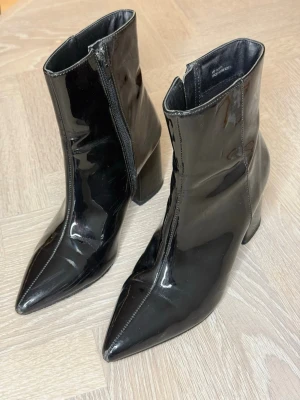 Svarta lackade stövletter med klack - Säljer ett par svarta stövletter i glansigt lackmaterial med spetsig tå och bred klack. Dragkedja på insidan för enkel på- och avtagning. Snygg retrovibe och passar perfekt till trendiga outfits.