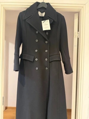 Svart lång dubbelknäppt kappa Zara - Stilren svart lång kappa från Zara med dubbelknäppning och bred krage. Kappan har två fickor framtill och markerad midja för en snygg siluett. Perfekt för dig som vill ha en klassisk och trendig look. Materialet är ullblandning.