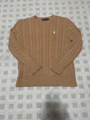 Beige Ralph Lauren tröja - Snygg beige kabelstickad tröja från Polo Ralph Lauren med klassisk vit broderad logga på bröstet. Tröjan har rund halsringning, långa ärmar och ribbade muddar. Perfekt för dig som gillar tidlös och stilren design. Inga tecken alls på användning!!