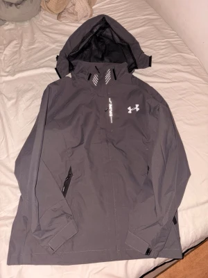 Grå vindjacka från Under Armour - Snygg grå vindjacka från Under Armour med huva och dragkedja. Jackan har vita logotyper och texten 'Explore' på bröstet. Tillverkad i ett lätt och vattenavvisande material, perfekt för blåsiga dagar. Justerbara muddar och fickor med dragkedja.