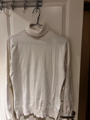 Vit polotröja i stickat material - Klassisk vit stickad polotröja med lång ärm och ribbade muddar. Tröjan har en hög polokrage och är tillverkad i ett tunt, mjukt material som passar perfekt för lager på lager.