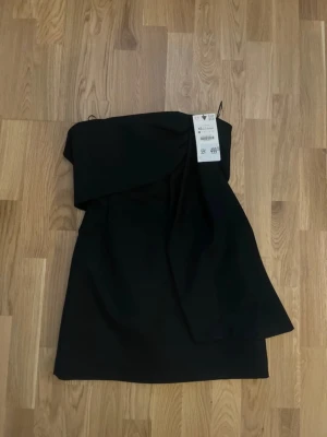 Svart axelbandslös kort klänning - Snygg svart axelbandslös kort klänning med asymmetrisk detalj framtill. Prislappen kvar! 