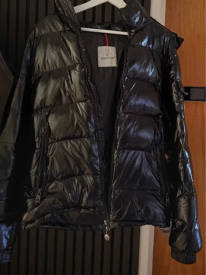 Svart pufferjacka från Moncler - Snygg svart pufferjacka från Moncler med glansig finish och klassisk logotyp på ärmen. Jackan har hög krage, dragkedja framtill och två sidofickor med dragkedja. Perfekt för kalla vinterdagar och riktigt trendig look.