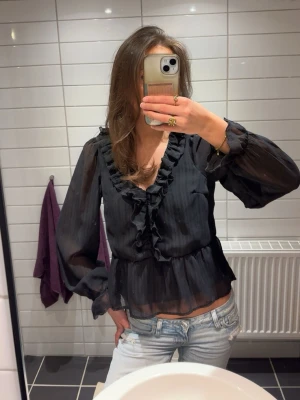 Svart volangblus  - Säljer en svart blus med volangdetaljer längs ringningen och framtill. Blusen har långa, transparenta ärmar och lätt utsvängd nederdel. Materialet är tunt och ger en luftig känsla. Perfekt för dig som gillar romantiska detaljer och en trendig look.