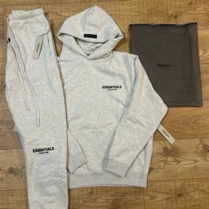 Ljusgrå Essentials hoodie - Säljer en ljusgrå hoodie från Essentials med svart broderad logga på bröstet. Hoodien har en klassisk huva och ribbade muddar vid ärmslut och nederkant. Perfekt för en avslappnad och trendig look. Priset är ej skrivit i sten och skrev för fler bilder.