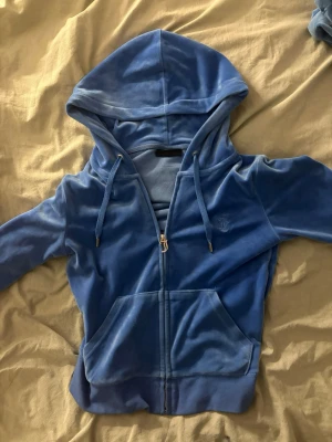 Blå velour hoodie med dragkedja - Snygg blå hoodie i mjuk velour från Juicy Couture. Hoodien har dragkedja framtill, två fickor och justerbar huva med snören. Perfekt för en chill och avslappnad stil. Liten logga broderad på bröstet.