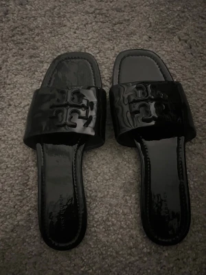 Svarta sandaler från Tory Burch - Snygga svarta sandaler från Tory Burch i glansigt lackmaterial. De har bred rem med det ikoniska Tory Burch-logotypen i relief och fyrkantig tå. Perfekta för en trendig och avslappnad stil.