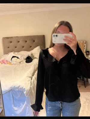 Svart blus med volangdetaljer - Snygg svart blus med långa ärmar och volangdetaljer längs knäppningen och kragen. Blusen har en lös passform och är tillverkad i ett tunt, lätt material som ger ett elegant intryck. Perfekt att styla med jeans eller kjol för en trendig look.