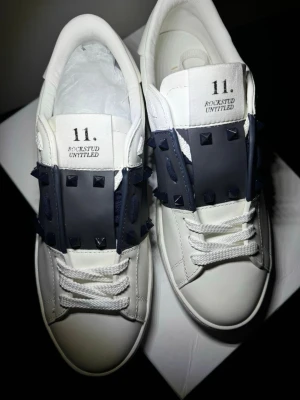 Valentino Rockstud Untitled sneakers - Snygga vita sneakers från Valentino med svarta nitar och en bred svart rem över snörningen. Skorna har en chunky sula med rutmönster undertill och är tillverkade i läder. Tungan har tryckt text med '11. ROCKSTUD UNTITLED' för en unik look.